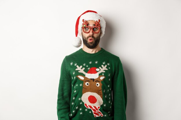 Christmas holidays, celebration concept. Surprised guy in party glasses and santa hat looking down amused, standing over white background