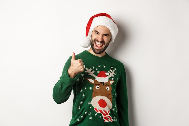 Christmas holidays, celebration and party concept. Man enjoying New Year, showing thumb up in approval, wearing Santa hat, white background.