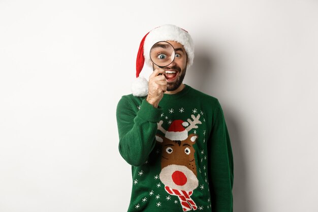 Concetto di natale e festività. un tipo barbuto divertente con un cappello da babbo natale che guarda attraverso la lente d'ingrandimento con stupore, ha trovato qualcosa, in piedi su uno sfondo bianco