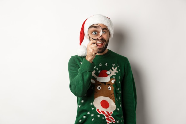 Концепция Рождества и праздников. Забавный бородатый парень в шляпе Санты с удивлением смотрит через увеличительное стекло, что-то нашел, стоя на белом фоне