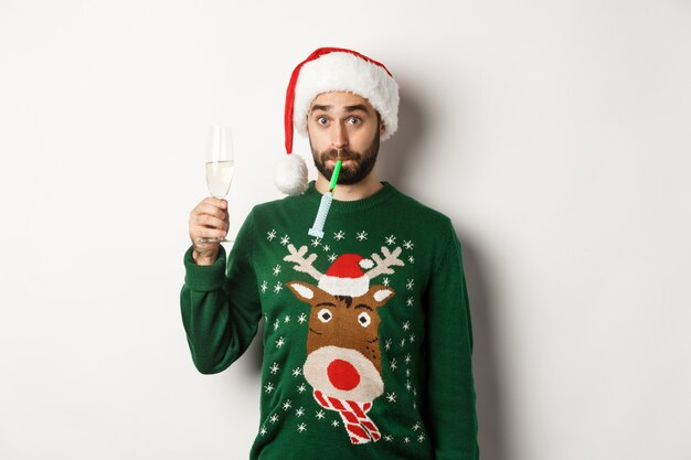Рождество и праздники концепция. Забавный парень в шляпе Санты, дует свисток, пьет шампанское, стоя на белом фоне