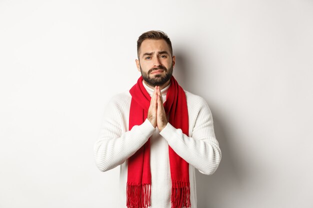 Foto gratuita vacanze di natale e concetto di capodanno. uomo disperato che implora aiuto, chiede favore, si tiene per mano in preghiera e guarda con speranza la telecamera, in piedi su sfondo bianco.