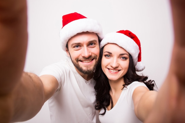 크리스마스, 휴일, 기술, 그리고 사람들의 개념 - 산타 모자를 쓴 행복한 커플이 흰색 바탕에 손에서 셀카 사진을 찍고 있습니다. 그는 그녀에게 키스한다.