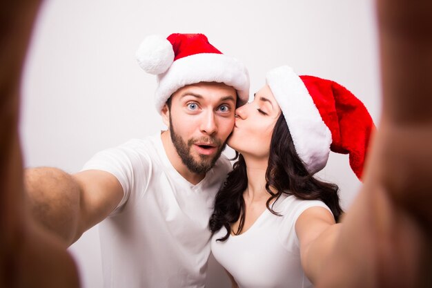크리스마스, 휴일, 기술, 그리고 사람들의 개념 - 산타 모자를 쓴 행복한 커플이 흰색 바탕에 손에서 셀카 사진을 찍고 있습니다. 그들은 카메라를보고 키스