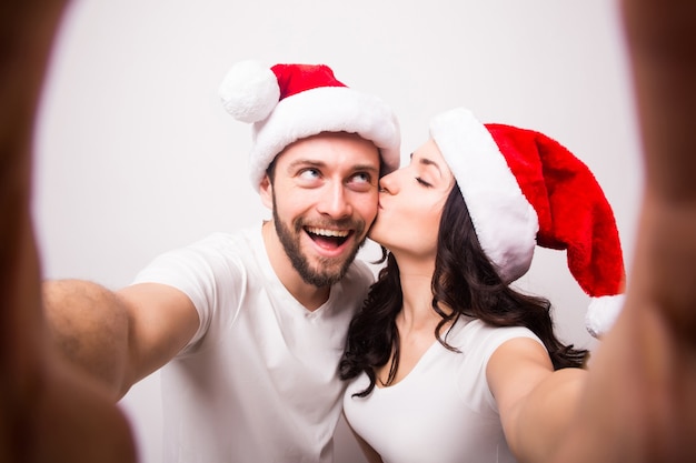 Christmas, holidays, technology and people concept - happy couple in santa hats taking selfie picture from hands on white background. They look at camera and kiss