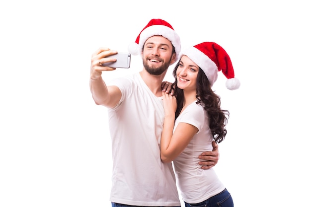 크리스마스, 휴일, 기술, 그리고 사람들의 개념 - 산타 모자를 쓴 행복한 커플이 흰색 바탕에 손에서 셀카 사진을 찍고 있습니다. 그들은 카메라를 찾습니다.