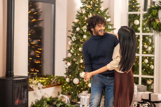 Christmas mood. Cheerful young curly indian man dancing paired with woman with long dark hair in lighted room with x-mas decoration