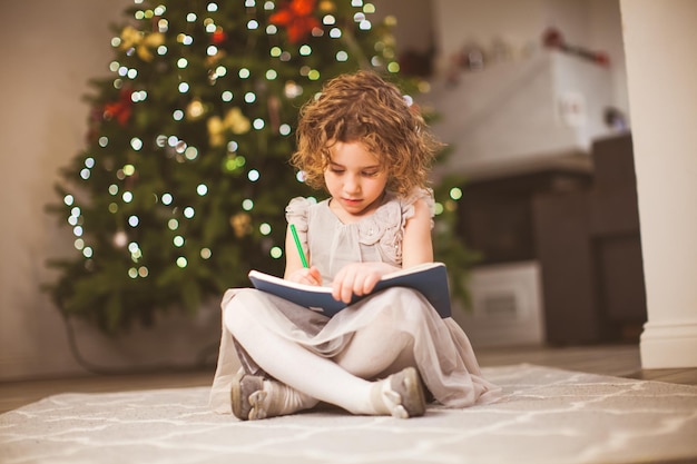 お祝いの木の横に座って、彼女のスケッチブックで描くかわいい巻き毛の女の子のクリスマス気分の写真家族の冬の休日のコンセプト Premium写真