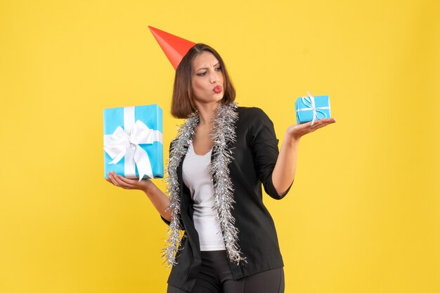 Рождественское настроение с эмоциональной бизнес-леди в костюме с рождественской шляпой, смотрящей на свои подарки на желтом