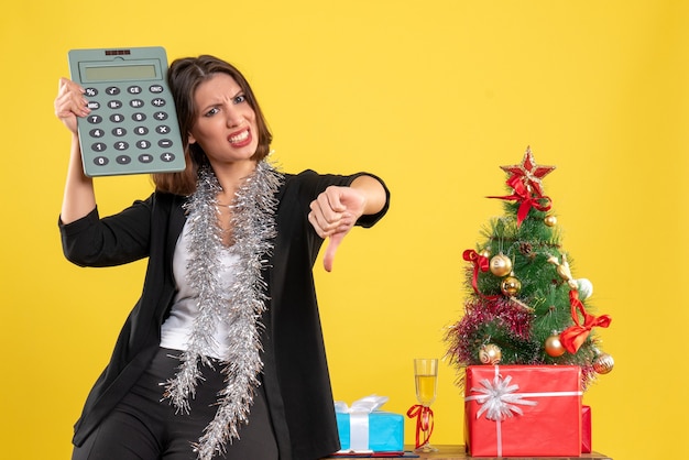 Christmas mood with nervous beautiful lady standing in the office and holding calculator making negative gesture in the office on yellow 