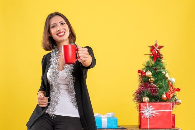 Christmas mood with positive beautiful lady standing in the office and holding red cup in the office on yellow 