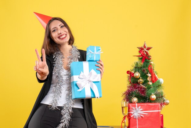 Christmas mood with positive beautiful lady with xsmas hat holding gifts with victory gesture in the office on yellow 