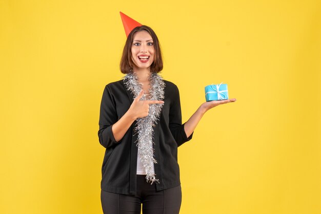 Christmas mood with positive business lady in suit with xsmas hat and pointing the gift on yellow 