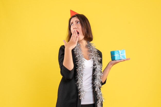 Christmas mood with shocked surprised business lady in suit with xsmas hat and holding gift on yellow 