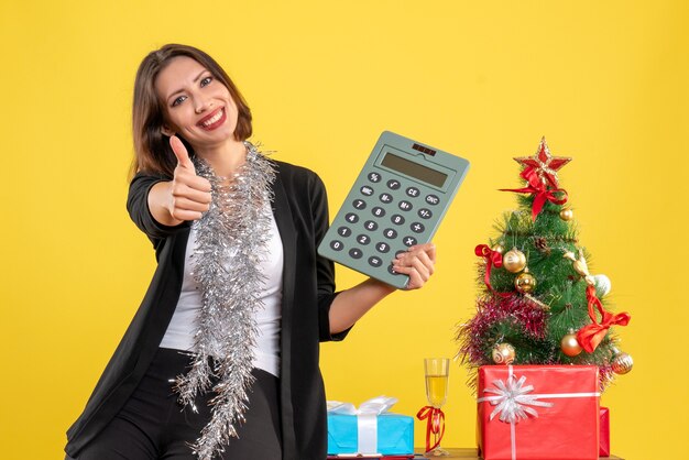Рождественское настроение с улыбающейся красивой леди, стоящей в офисе и указывающей на калькулятор, делая жест в офисе на желтом