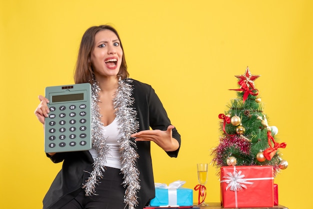 オフィスに立っている笑顔の美しい女性と黄色のオフィスで電卓を指すクリスマス気分