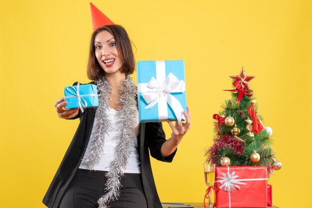 Atmosfera natalizia con bella signora sorpresa con cappello di natale che mostra felicemente i regali in ufficio su giallo