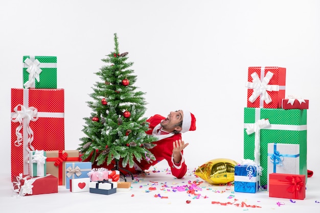 Atmosfera natalizia con babbo natale sorpreso che si nasconde dietro l'albero di natale vicino a regali in diversi colori su sfondo bianco