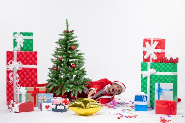 Новогоднее настроение с молодым грустным дедом морозом, лежащим за елкой возле подарков разных цветов на белом фоне