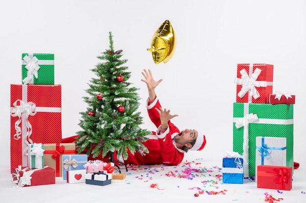 白い背景画像にさまざまな色の贈り物の近くのクリスマスツリーの後ろに横たわっている若いサンタクロースとクリスマス気分