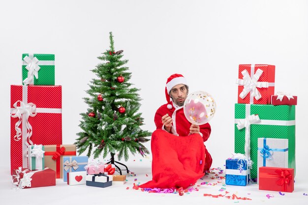 흰색 배경에 다른 색상의 크리스마스 트리와 선물 근처에 앉아 젊은 산타 클로스와 크리스마스 분위기