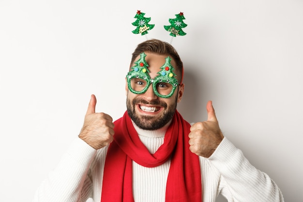 Christmas, New Year and celebration concept. Close-up of handsome bearded man in funny party glasses looking happy, showing thumb up in approval, like something, white background