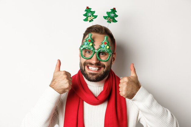 Christmas, New Year and celebration concept. Close-up of handsome bearded man in funny party glasses looking happy, showing thumb up in approval, like something, white background