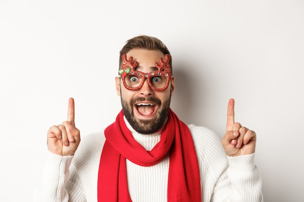 Christmas, New Year and celebration concept. Close-up of handsome bearded man in party glasses and red scarf, celebrate winter holidays and pointing fingers up at copy space