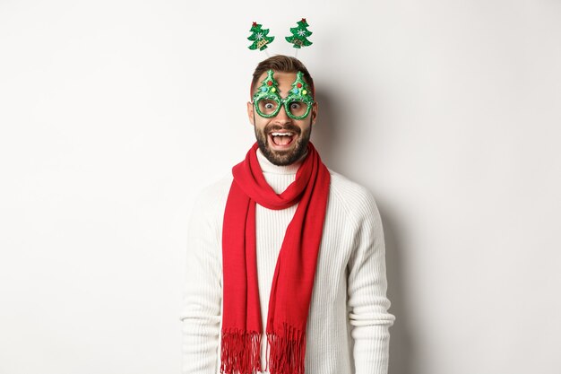 Christmas, New Year and celebration concept. Handsome bearded man looking surprised, wearing party glasses and accessory, white background