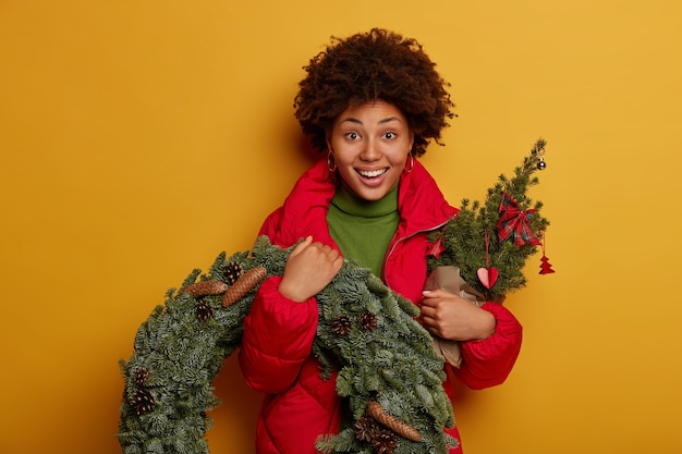 Christmas and New Year concept. Curly haired young woman carries fir tree and small wreath, gets ready for winter holidays, wears red coat