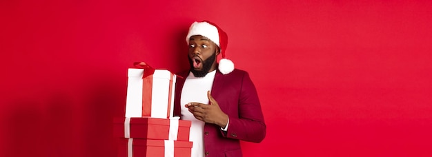 Foto gratuita natale, capodanno e concetto di shopping hanno sorpreso l'uomo di colore guardando i regali di natale con stupore