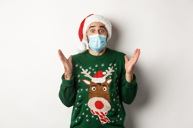 Рождество во время пандемии, концепция covid-19. Удивленный парень в медицинской маске, шляпе Санты и свитере празднует новогоднюю вечеринку, стоя на белом фоне