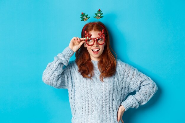 Christmas party and celebration concept. Cute redhead teen girl celebrating New Year, wearing xmas tree headband and funny glasses, looking left amused, blue background.