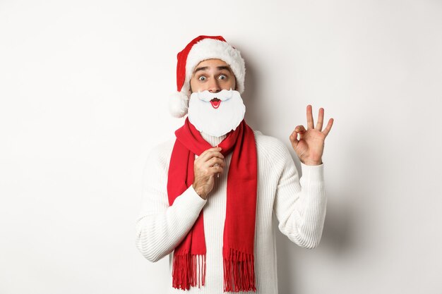 크리스마스 파티와 축 하 개념입니다. 흰색 배경 위에 서 있는 확인 제스처를 보여주는 산타클로스 모자와 흰 수염 마스크를 쓴 행복한 남성 모델