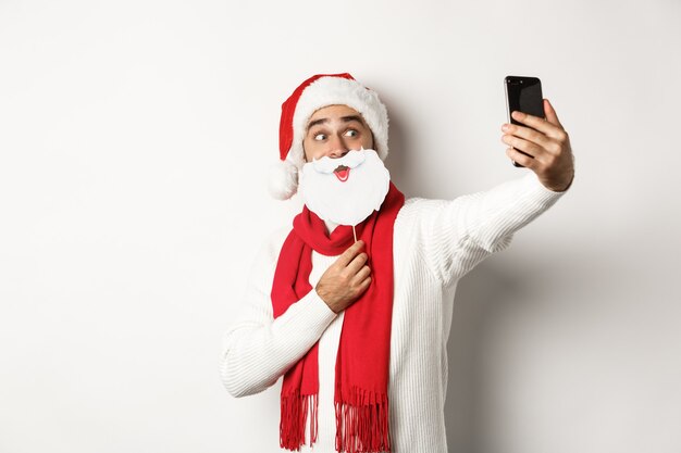Christmas party and celebration concept. Young man taking selfie with funny white beard Santa mask and hat, posing for photo on mobile phone, studio background