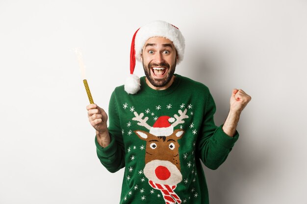 Рождественская вечеринка и концепция праздников. Красивый бородатый мужчина радуясь, держа фейерверк бенгальского огня и нося шляпу Санты, празднуя Новый год, белый фон.