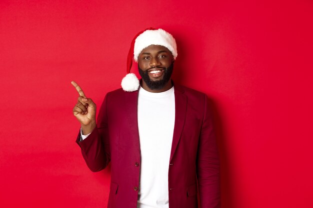 クリスマス、パーティー、休日のコンセプト。左指を指して、広告を表示し、赤い背景の上に幸せに立っているサンタ帽子のハンサムな黒人男性