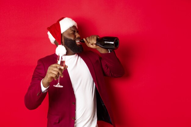 Рождество, вечеринка и праздничное понятие. Изображение молодого темнокожего человека в новогодней шапке, пьющего шампанское из бутылки, напивающегося на праздновании Нового года, стоящего на красном фоне