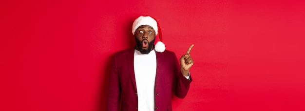 Christmas party and holidays concept impressed black man with beard wearing santa hat pointing finge