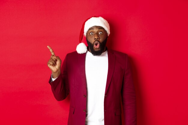 크리스마스, 파티 및 휴일 개념입니다. 수염을 기른 흑인 남자, 산타 모자를 쓰고 손가락을 왼쪽으로 가리키고 놀란 듯 헐떡이며 빨간색 배경에 서서