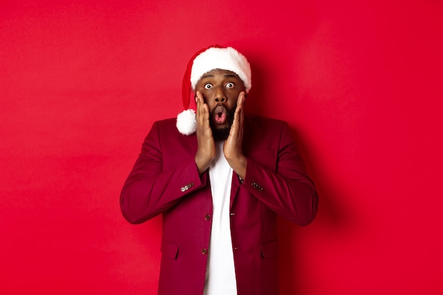 Christmas, party and holidays concept. Shocked Black man in santa hat reacting to new year offer, standing against red background.