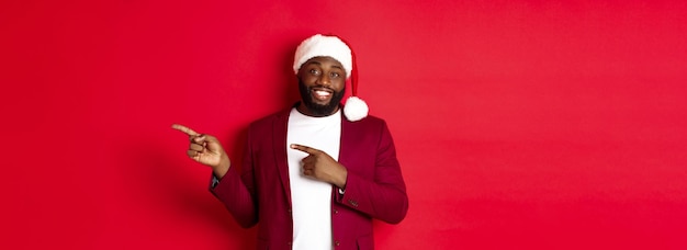 Free photo christmas party and holidays concept smiling black man with beard and santa hat pointing fingers rig