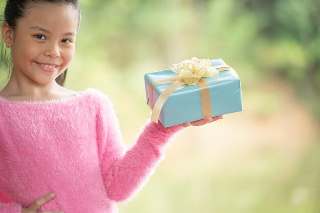 녹색 분기 나무 근처 선물 상자와 함께 행복 하 게 웃는 어린 여자 아이의 크리스마스 초상화. 녹색 자연 숲에서 초점 배경 bokeh 나뭇잎.