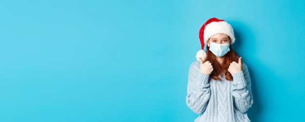 Foto gratuita quarantena natalizia e concetto covid cute teen redhead ragazza in cappello di babbo natale e maglione che indossa il viso