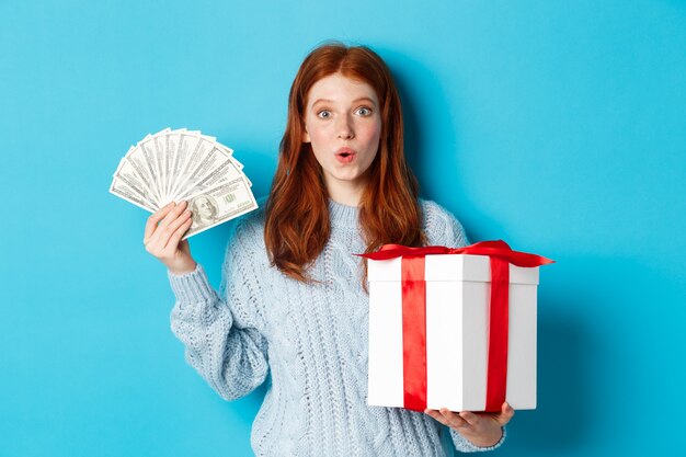 Рождество и концепция покупок. Возбужденная рыжая девушка смотрит в камеру, держит большой новогодний подарок и доллары, покупает подарки, стоя на синем фоне.