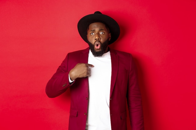 Christmas shopping and people concept. Worried african american man pointing at himself, being accused, standing nervous against red background