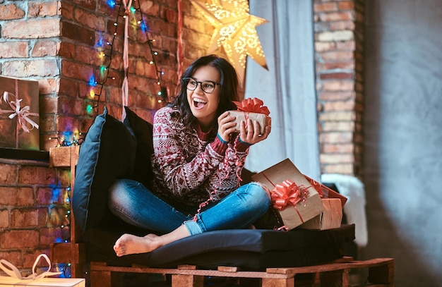 Natale, san valentino, capodanno. bella ragazza che si gode la mattina di natale con in mano una confezione regalo mentre è seduto su un divano in una stanza decorata con interni soppalcati.