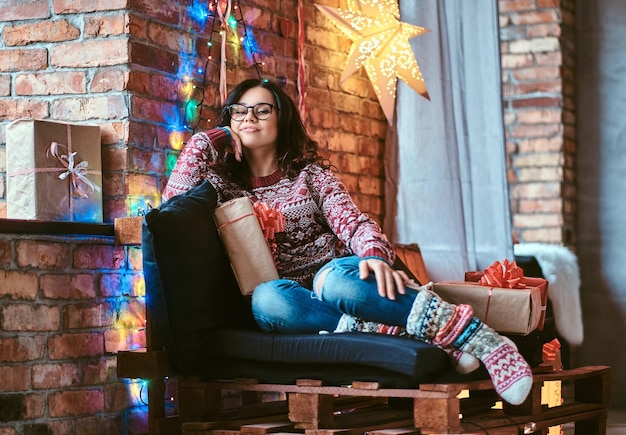 Christmas, Valentine's Day, New year. Happy beautiful girl enjoying Christmas morning while sitting on a couch with gift boxes in a decorated room with loft interior.
