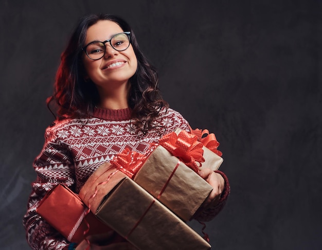 Natale, san valentino, capodanno. ritratto di una ragazza bruna felice che indossa occhiali da vista e maglione caldo che tiene scatole di regali, isolato su uno sfondo scuro con texture.