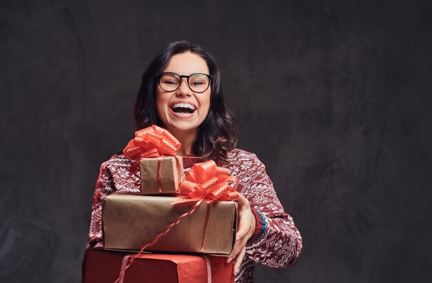 クリスマス、バレンタインデー、正月。暗いテクスチャの背景に分離された、ギフトボックスを保持している眼鏡と暖かいセーターを着ている幸せなブルネットの少女の肖像画。